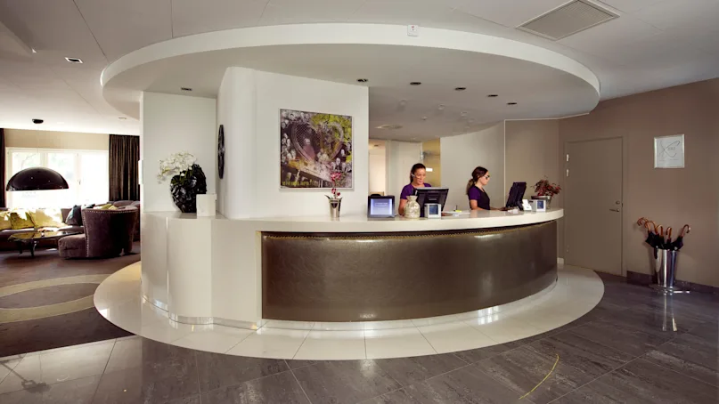 Two staff members work at a curved reception desk in a modern hotel lobby.