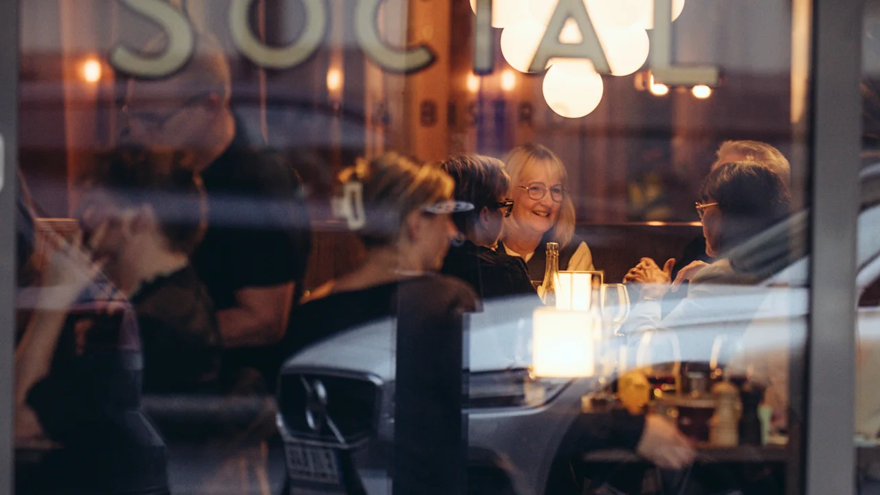 En grupp människor sitter inne på ett varmt upplyst café, sett genom ett fönster med ordet "Social" på, medan de pratar och ler runt ett bord. En bil speglas i glaset.