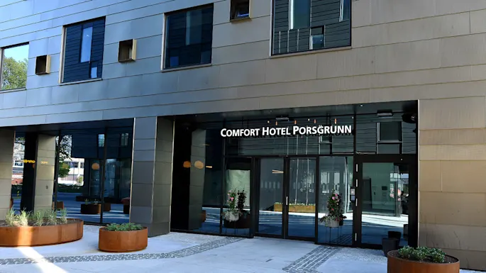 Entrance with glass doors displaying "COMFORT HOTEL PORSGRUNN" in urban setting, surrounded by large planters with green plants and modern architecture.