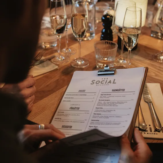 A person holding a menu labeled "The Social Bar & Bistro," seated at a table set with wine glasses, utensils, and a lit candle in a restaurant environment.
