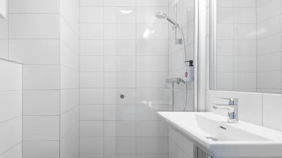 Dusch med krombeslag i ett kaklat badrum; vit tvättställ med vägghängt spegel, stark belysning ovanför och flytande tvåldispenser på väggen.