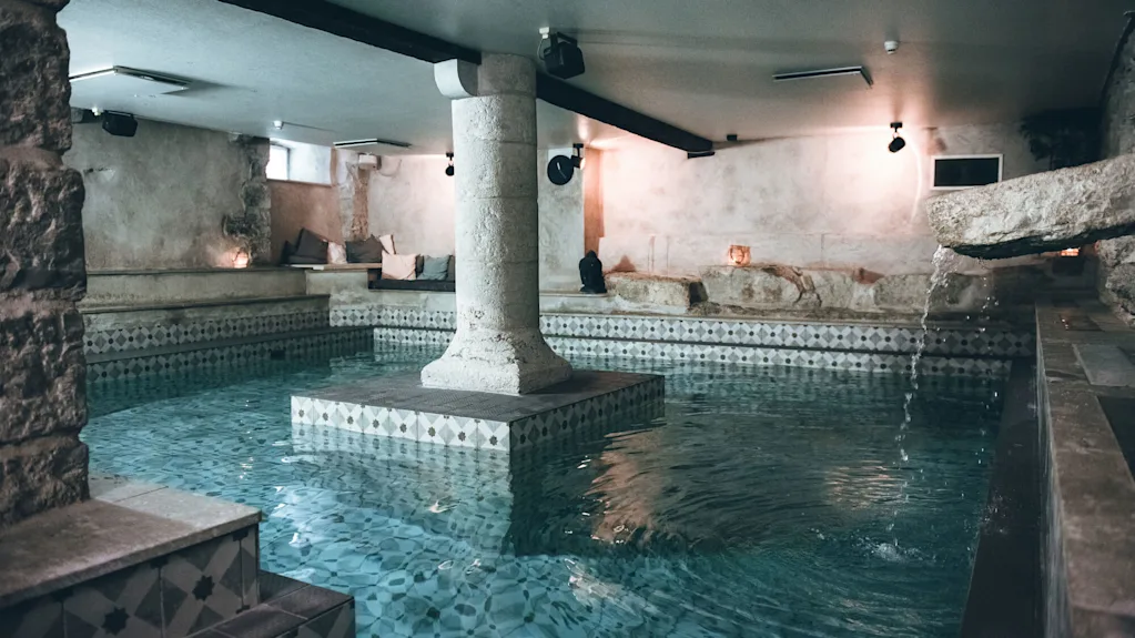 A stone column rises from a serene, tile-patterned indoor pool, surrounded by soft lighting and stone walls, with water gently cascading from a spout.
