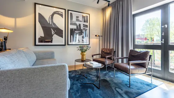 A modern living room with a gray sofa, leather armchairs, abstract wall art, and a large window that lets in daylight.