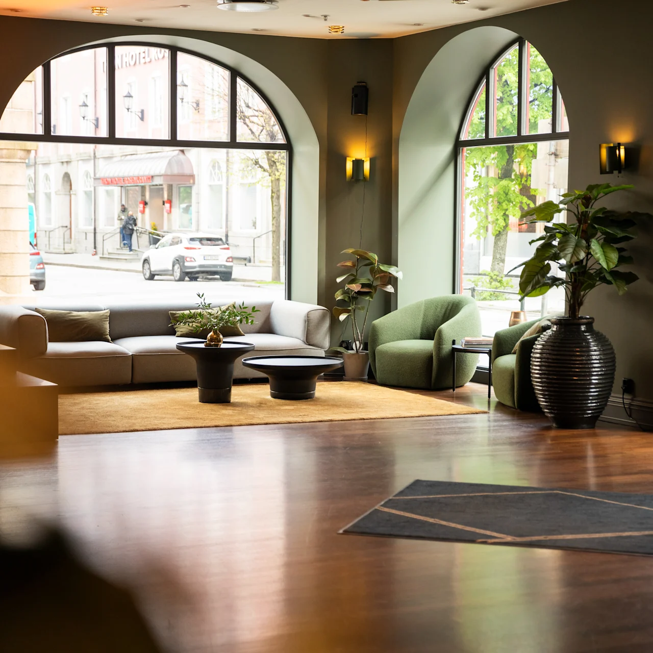 Et siddeområde med en moderne grå sofa og en grøn lænestol står i et rum med store buede vinduer, der vender ud mod en gade. Potteplanter tilføjer grønt til indretningen.