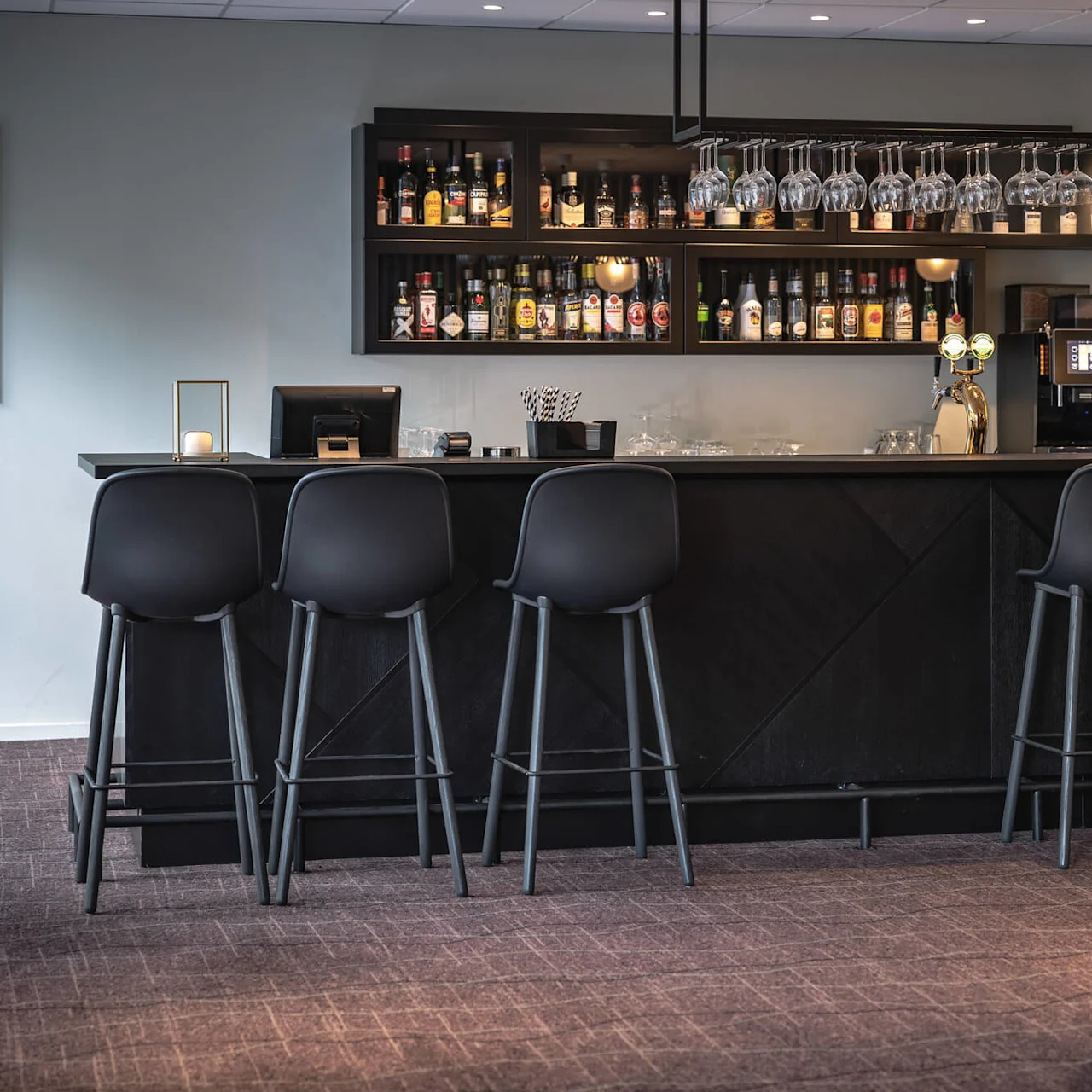 En moderne bar med seks sorte stole vendt mod en disk; hylder bagved viser forskellige spiritusflasker, omgivet af hængende glas. Blød belysning skaber en hyggelig atmosfære i loungen.