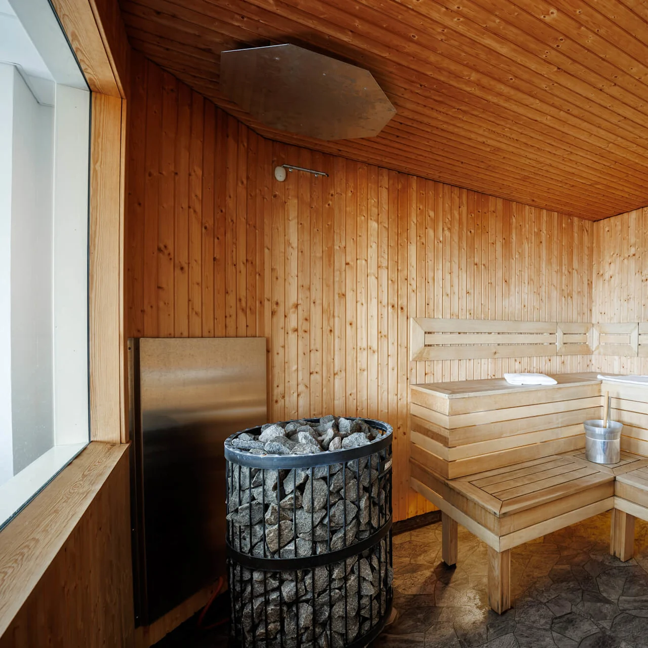 Ett träbeklätt basturum har en hörnbänk med en handduk och hink, en stenugn som avger värme, och en väggtermometer med texten "SAUNA" vilket skapar en mysig miljö.