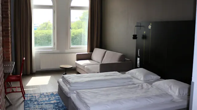 Två prydligt bäddade enkelsängar vilar på en mönstrad blå matta i ett ljust rum, med en grå soffa nära stora fönster och ett litet rött skrivbord i hörnet.