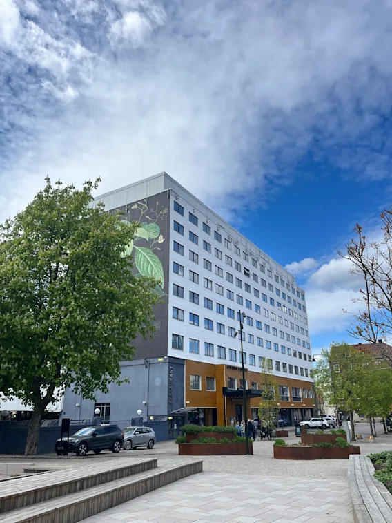 Byggnad med lövmålning står vid trädkantad gata under delvis molnig blå himmel. Bilar och fotgängare syns.