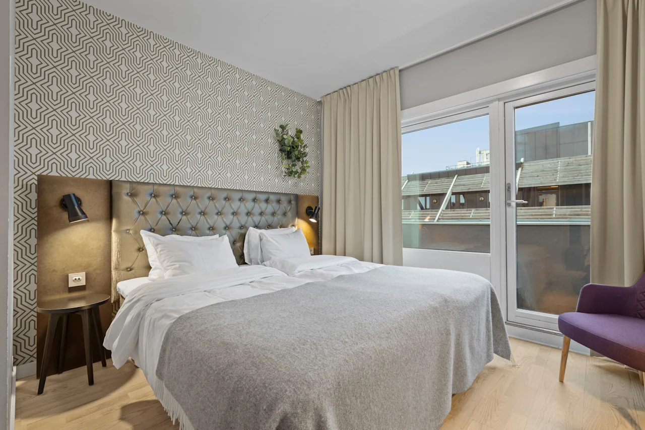 Ett mysigt hotellrum med en stor säng, mönstrad tapet och ett fönster med utsikt över närliggande byggnader. En lila stol står vid fönstret.