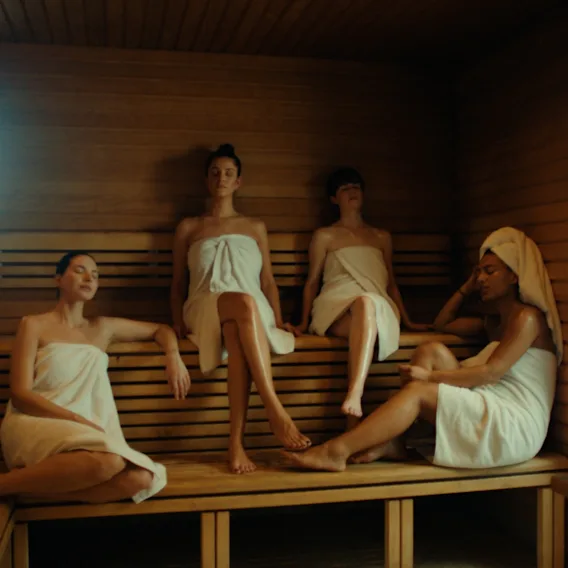 Four people wrapped in towels relax on wooden benches in a sauna, one resting their head with a towel wrap. Sunlight filters through a small window, enhancing the serene ambiance.