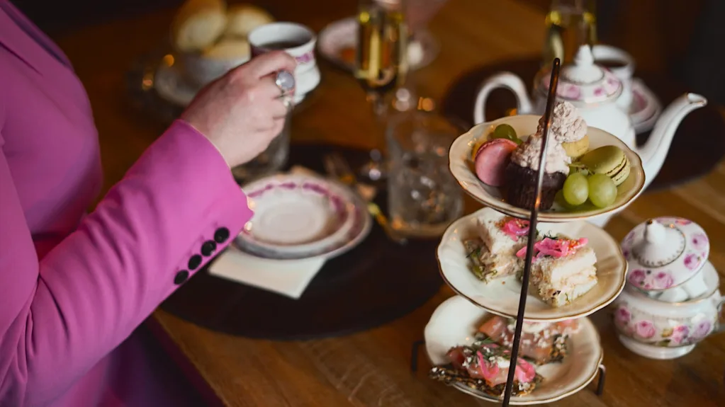 En person i en pink jakke nyder eftermiddagste med et udvalg af kager på et etagers fad, elegant porcelæn og en kop te, indendørs.