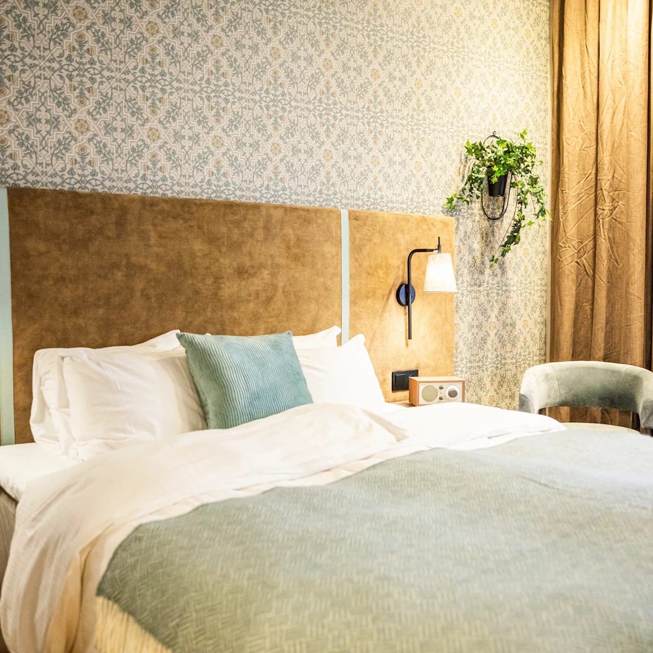 A neatly made double bed with white and blue bedding rests against a patterned wall. To the right, a chair and desk with stacked books sit near closed brown curtains.