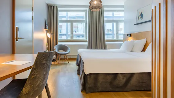 A modern hotel room with a large bed, wooden floors, desk and chair, under soft lighting, facing windows with city view.