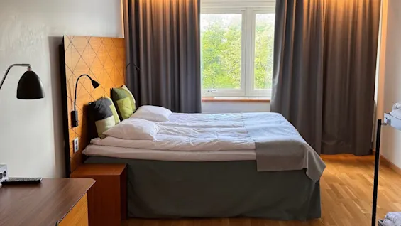 Bed with white linens and cushions positioned beside a wooden desk and lamp, set in a room with wooden flooring. Large window with gray curtains reveals greenery outside.