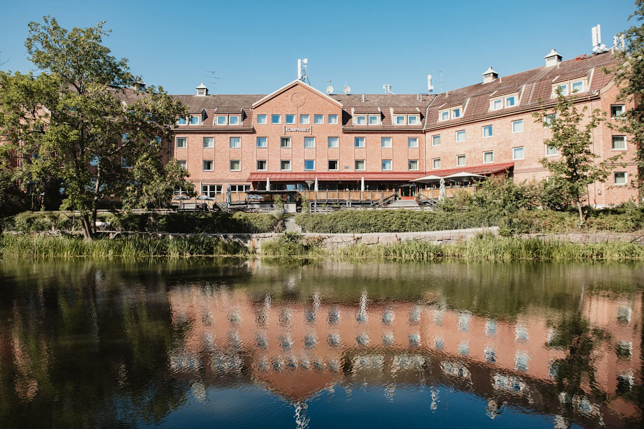 En stor tegelbyggnad med en skylt där det står KOMPANIET speglas i ett lugnt vattendrag med träd och grönska.