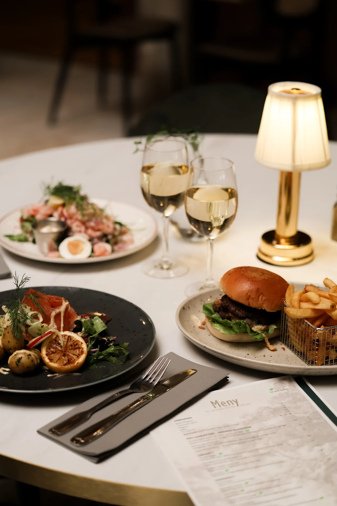 Burger with fries sits on a table next to a salad, two glasses of white wine, a plate with smoked salmon and potatoes, a menu, and a small lamp. 