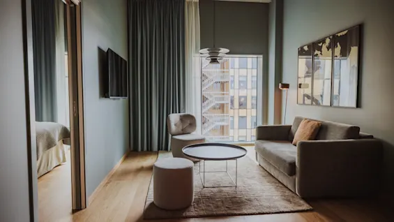Mysig vardagsrum med grå soffa och stol runt ett runt glasbord. En matta täcker trägolvet. Väggkonst och ett gardinbeklätt fönster skapar en lugn, modern atmosfär.
