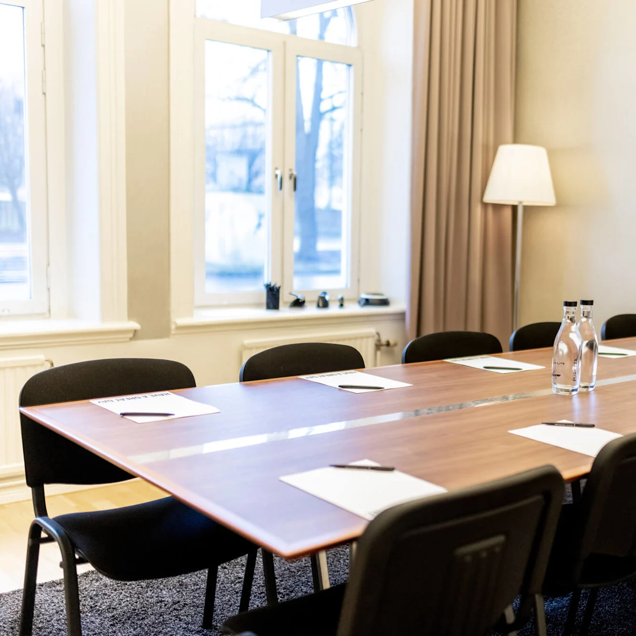 A conference room with a large table, chairs, papers, and water bottles, ready for a meeting. There's a TV and a closed curtain.