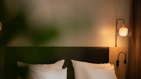 Bed with white pillows rests against a quilted headboard, softly lit by a wall-mounted lamp. The setting appears cozy and relaxing with an ambient glow in the room.