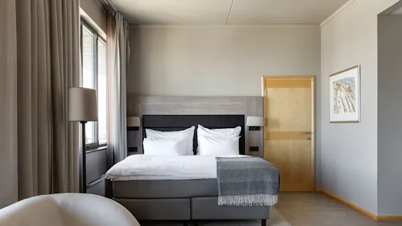A neatly made bed is in a modern room, flanked by floor-length curtains and a lamp, with a pale wooden door and framed art on the wall.
