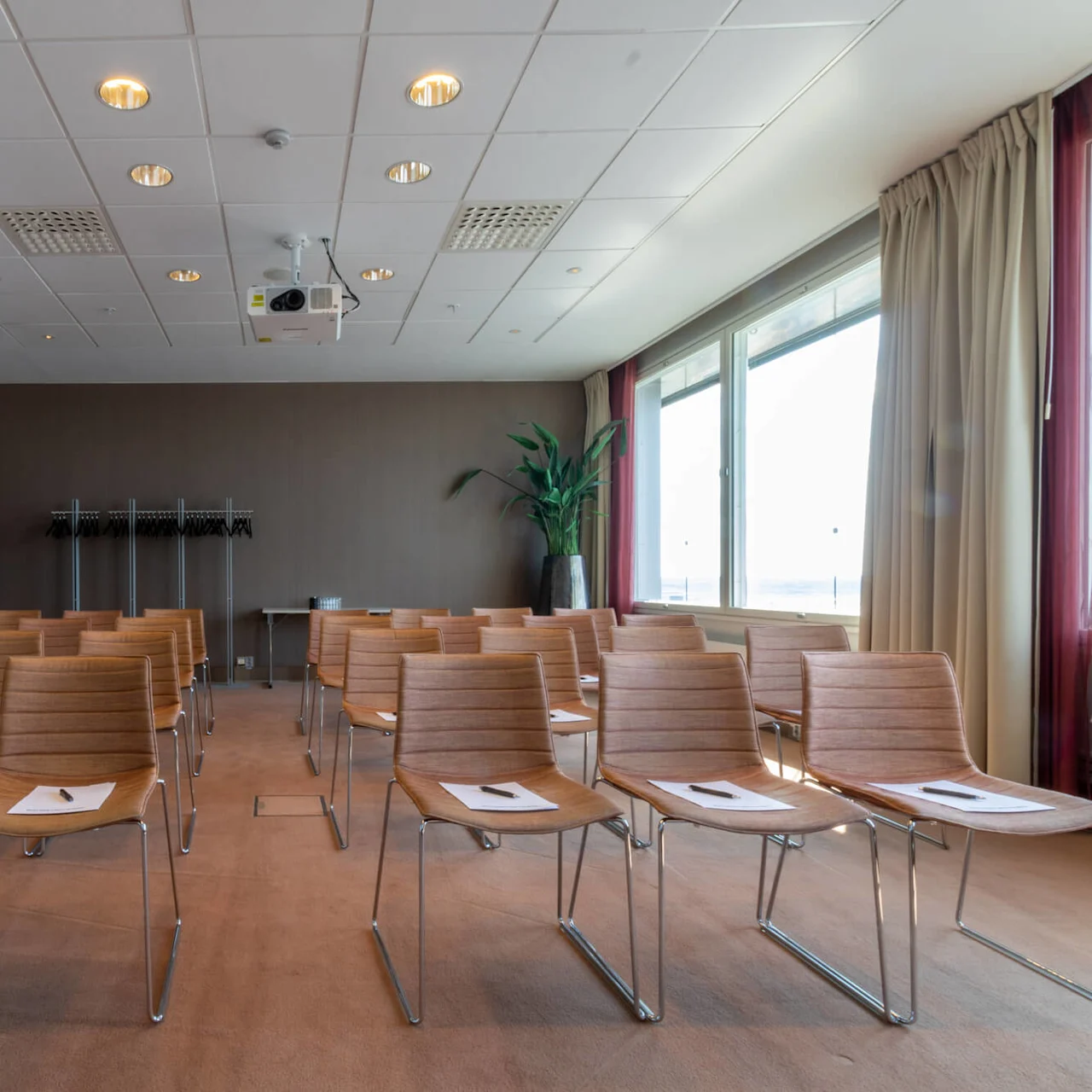 Rows of brown chairs face forward, each with a paper and pen, in a conference room. A coat rack and large plant are in the background. A window with curtains is on the right.