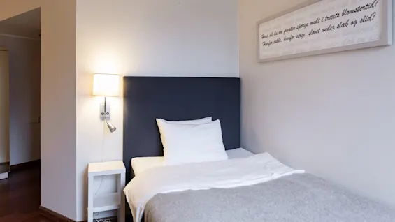 Ett hotellrum med en enkelsäng, lampa och en inramad citat: Hvad vil du om frugten spørge midt i træets blomstertid?