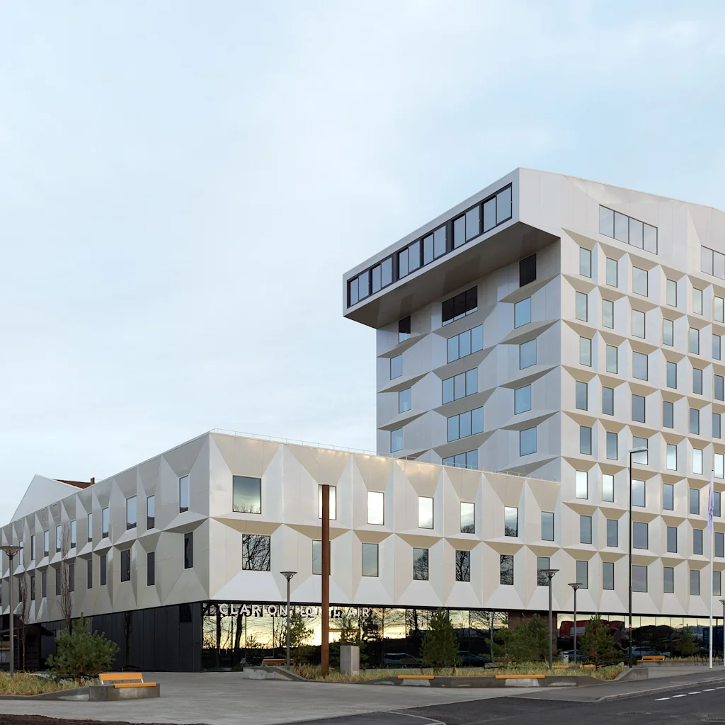 En moderne hotelbygning med en geometrisk facade og store vinduer. Et fly flyver på himlen ovenover. Synlig tekst: HOTEL