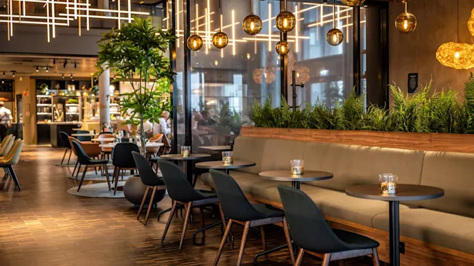 Elegant seating area features green upholstered chairs and wooden tables; ambient lighting and indoor plants create a cozy, stylish atmosphere in a modern restaurant setting.