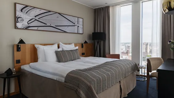 Et moderne hotellrom med en komfortabel seng, store vinduer med utsikt over byen og moderne innredning.