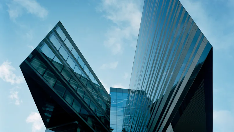 A modern, angular glass building extends upward, reflecting the blue sky with scattered clouds. The architectural design features sharp, intersecting lines creating a dynamic urban aesthetic.