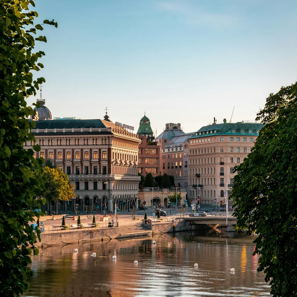 En naturskön vy över Stockholm med historiska byggnader längs en lugn flod, inramad av gröna träd och ett utomhuskafé.