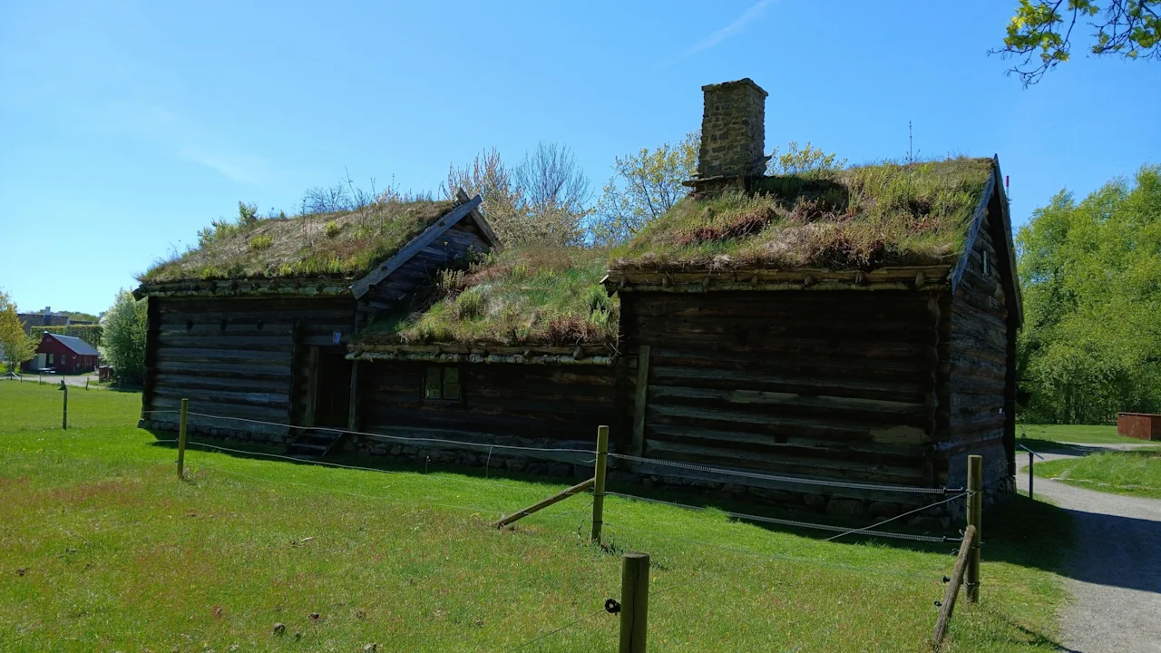 Fredriksdal museer och trädgårdar: Tradisjonelle trebygninger med grønne gresstak under en klarblå himmel, omgitt av natur.