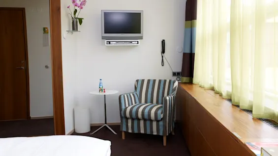 A striped armchair sits beside a small round table with bottled water and toys in a hotel room with a wall-mounted TV, orchid, and large curtain-covered window.