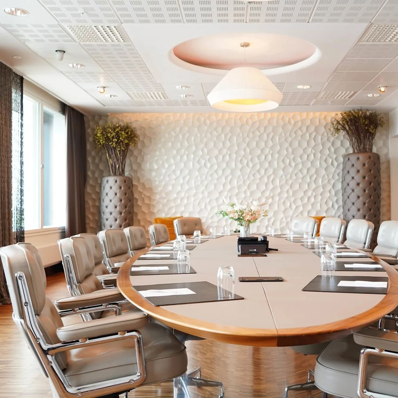A large oval conference table surrounded by leather chairs, set in a modern room with textured walls, large planters, and windows with curtains. Papers and glasses are on the table.
