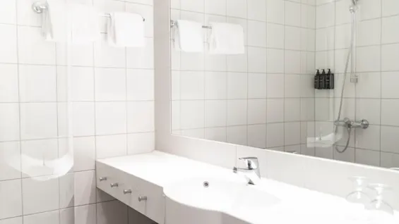 Et lyst, moderne hotellbad med hvite fliser, et stort speil, en vask og et dusjområde med rene håndklær.