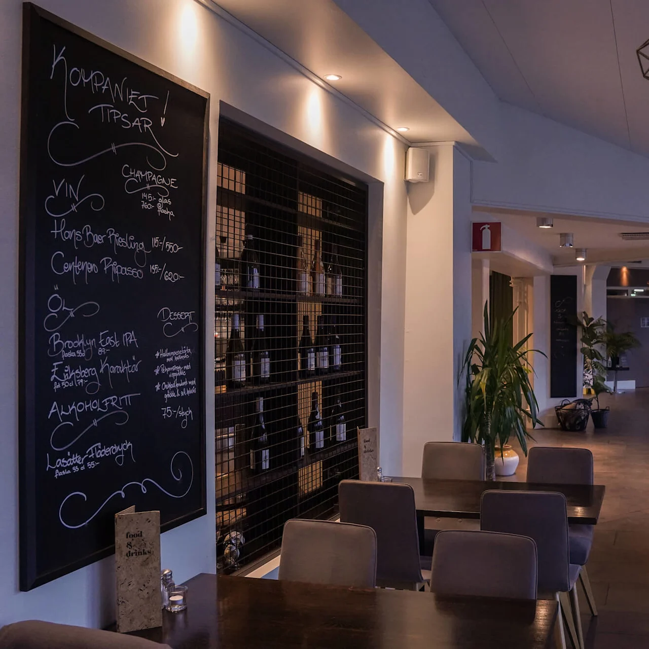 A dimly lit restaurant interior with chairs around tables; a chalkboard menu is visible on the left.