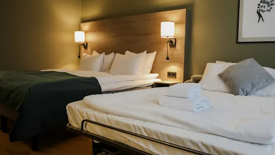 A neatly arranged hotel room features two beds, one with a green blanket and plush pillows, and a smaller bed with folded towels; wooden bedside tables and framed artwork enhance the cozy ambiance.