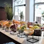 A buffet table presents a variety of colorful fruits and cheeses, neatly arranged in bowls and stands, set in a bright room with large windows and decorative plants. 