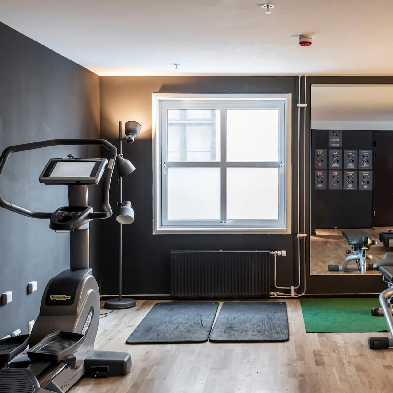 Träningscykel står stilla; spegelvägg, bänk, kettlebells och ihoprullade mattor organiserade intill. Ljust, trägolv i gym med naturligt ljus från stort fönster.