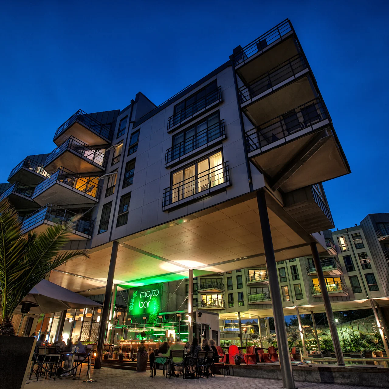 En fleretasjes moderne bygning med opplyste balkonger ser ut over et livlig utendørs barområde. Neonskiltet sier "mojito bar" mot en klar kveldshimmel.