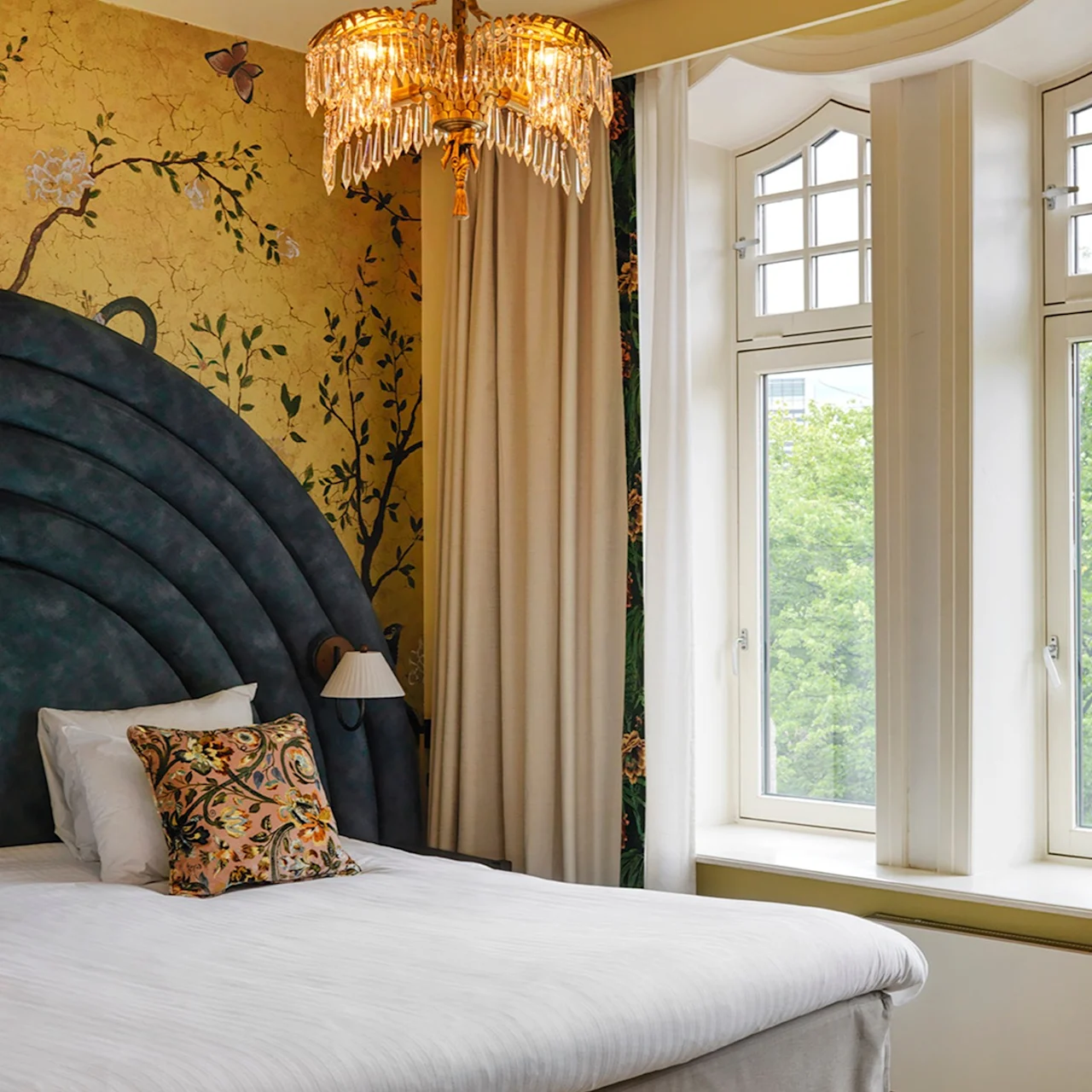 A bed is placed against a floral wallpaper and large arched headboard, adorned with decorative pillows. A chandelier hangs above, and a bay window offers a view of greenery outside.