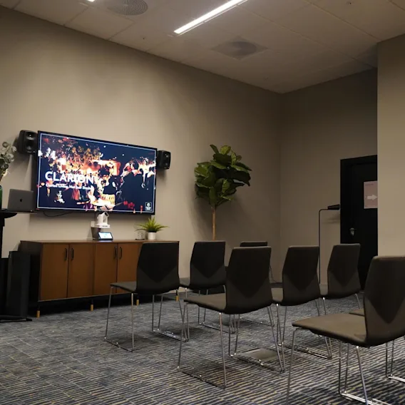 A screen showing "Clarion" above rows of chairs. A cabinet with speakers and decorations sits underneath. A potted plant is beside the door in a modern conference room.