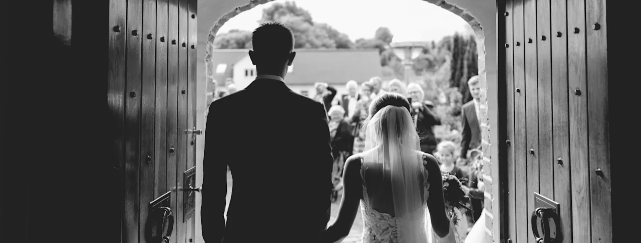 Ett par i formell klädsel går hand i hand genom en välvd dörröppning mot en väntande folkmassa.