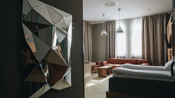 A geometric mirror reflects fragmented shapes in a modern room. The space includes a leather sofa, wooden tables, and large windows with curtains, giving a cozy and stylish ambiance.