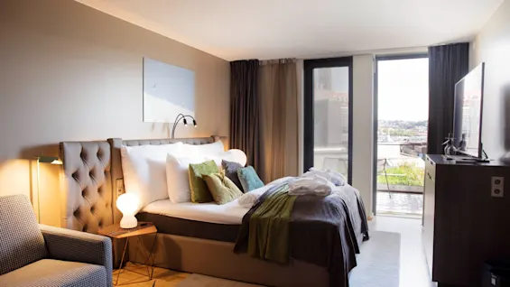 A bed in a hotel room, neatly made with pillows and throws, faces a large window. The room is warmly lit and furnished with a chair and a television.