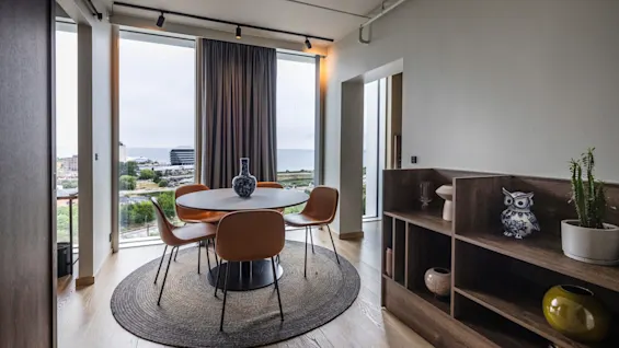 Et rundt bord med en vase er omgitt av fire brune stoler, med utsikt over et bylandskap gjennom store vinduer. Bokhyller med dekorasjoner linjerer den tilstøtende veggen.