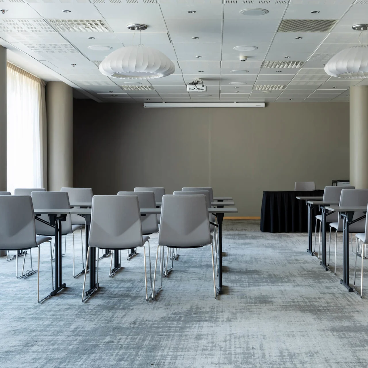 Rows of grey chairs face a blank wall under modern light fixtures in a spacious conference room with large windows and carpeted flooring.