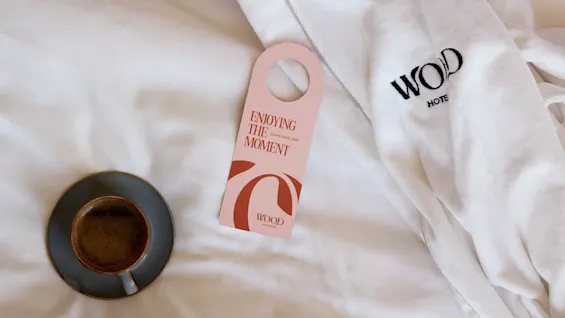 A coffee cup sits on a bed beside a robe labeled "WOOD Hotel" and a door hanger reading "ENJOYING THE MOMENT Come back later."