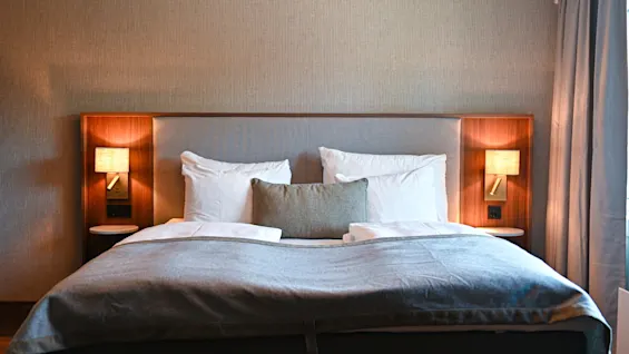 Et hyggeligt hotelværelse med en redt seng, bløde puder og varm sengelampebelysning, der indbyder til afslapning.