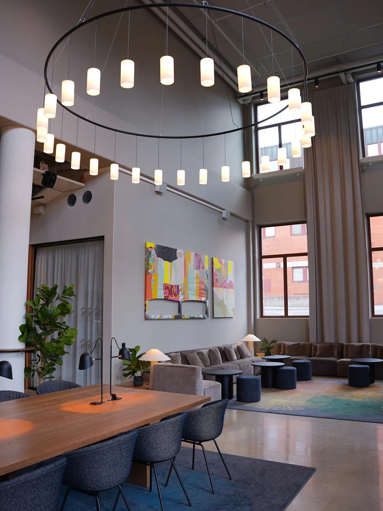 En hemtrevlig hotellounge med en stor cirkulär ljuskrona, modern konst på väggarna, mjuka soffor och ett långt träbord med blå stolar. Höga fönster släpper in naturligt ljus och skapar en inbjudande atmosfär.